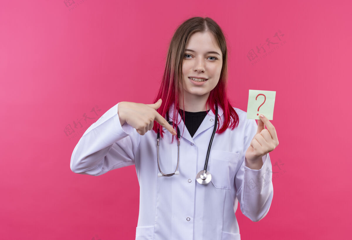 女孩穿着听诊器医用长袍的年轻医生女孩高兴地指着她手上粉红色孤立背景上的纸问号医学粉红色问题