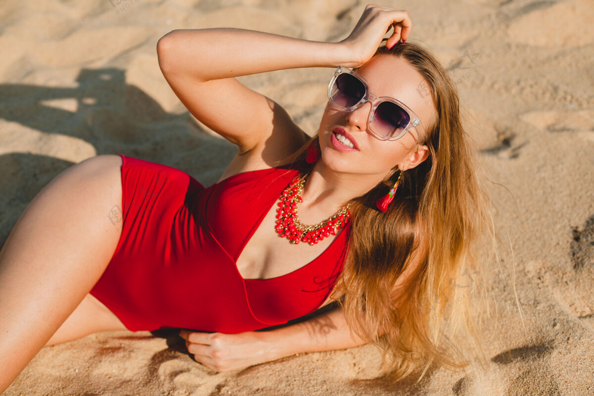 皮肤年轻漂亮的金发女人穿着红色泳衣 戴着墨镜在沙滩上晒太阳肖像身体乐趣