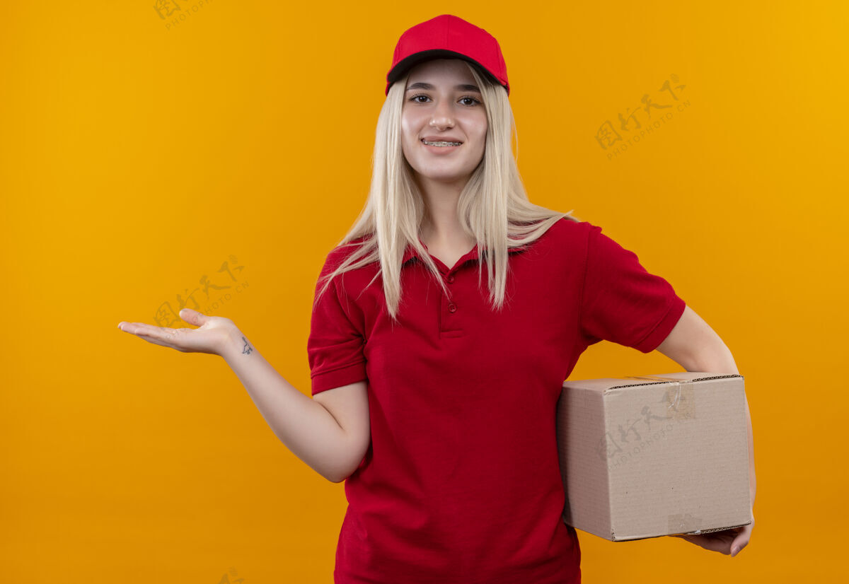 送货带着微笑的小女孩穿着红色t恤 戴着帽子 戴着牙套 站在一个孤立的橙色背景上微笑支架年轻