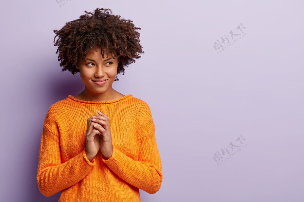 衣服高兴的非裔美国妇女制定计划 有耐人寻味的看旁边的自由空间 保持手掌在一起 看向别处 穿橙色套头衫 孤立在紫色的墙壁人 意图概念休闲热情手势