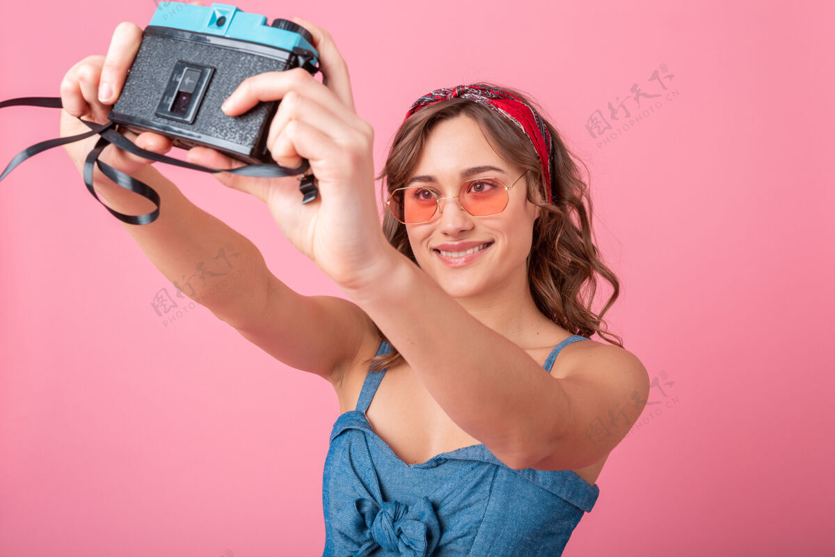 女孩迷人的微笑女人在老式相机上拍摄自画像 穿着牛仔裙 戴着粉色背景的太阳镜漂亮粉色带着