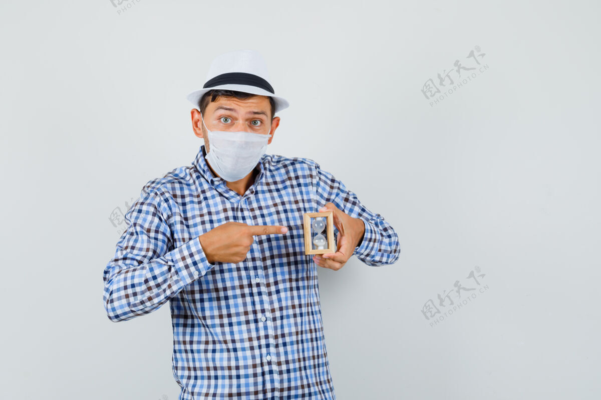 人一个穿着格子衬衫 戴着帽子 戴着面具的年轻人指着沙漏 看上去很担心人模特相机