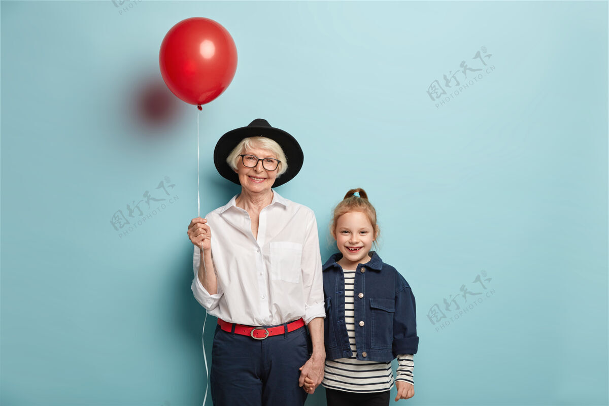 头饰微笑的红发女孩带着母亲节来祝贺奶奶 穿着条纹套头衫和牛仔夹克高兴的老太太戴着时髦的黑色帽子 拎着气球 牵着小孙女的手时尚奶奶奶奶