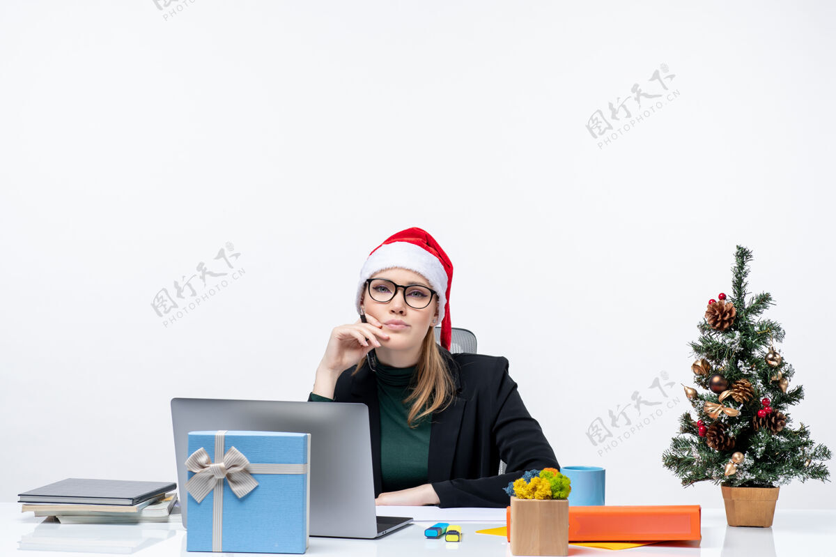 笔记本电脑一个体贴的金发女人 戴着圣诞老人的帽子 坐在一张桌子旁 桌子上放着一棵圣诞树和一份白色背景的礼物桌子帽子圣诞老人