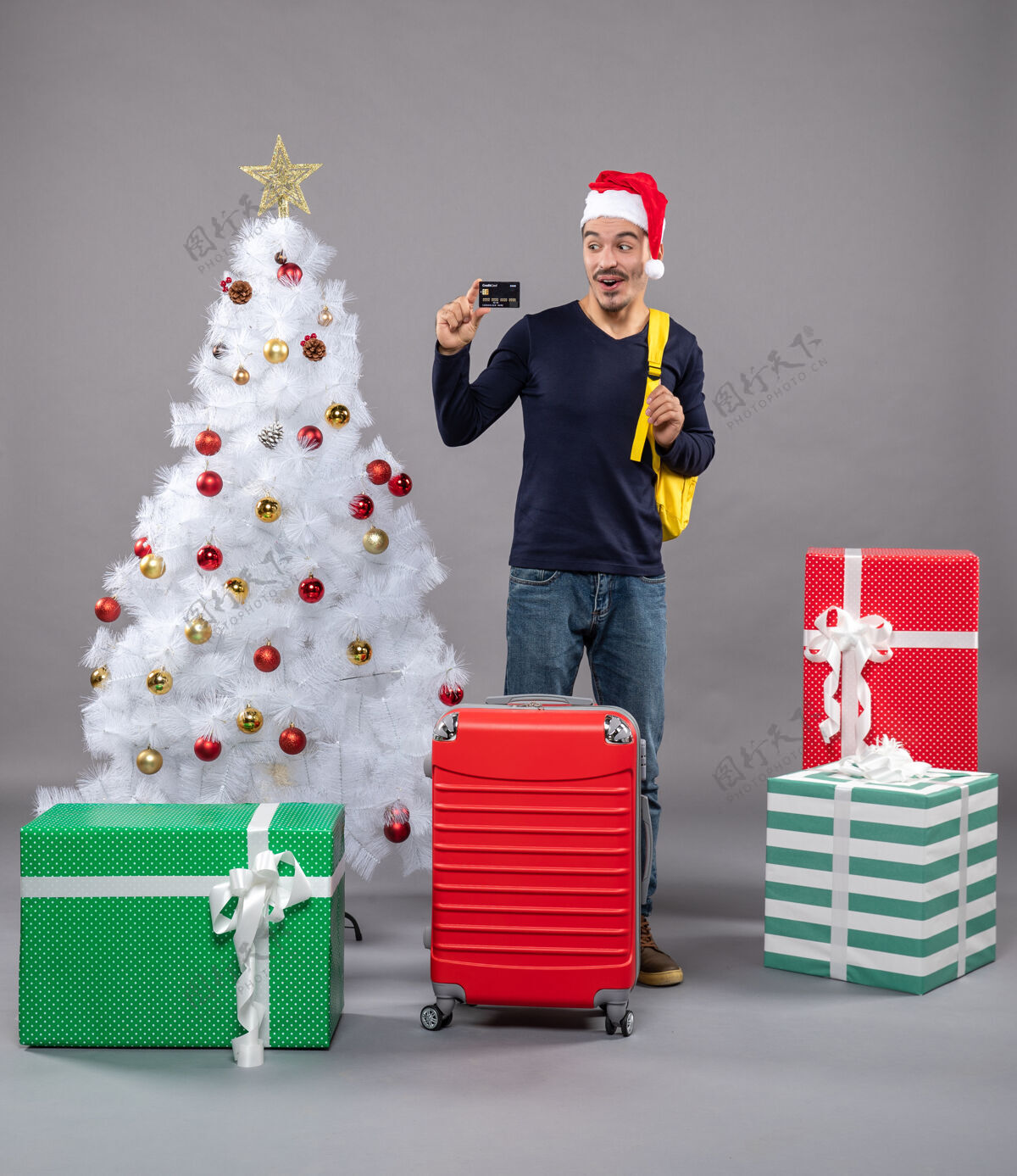 圣诞老人兴奋的年轻人拿着卡片站在灰色的圣诞树旁礼物包礼物