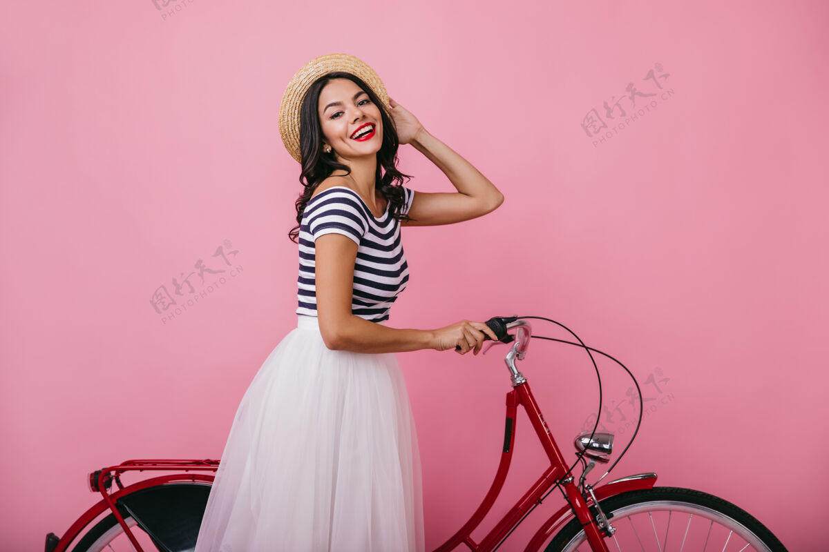 乐趣晒得黝黑的幸福女士戴着帽子玩着自行车穿着郁郁葱葱的裙子站着的幽默女孩的室内画像卷发自行车手帽子