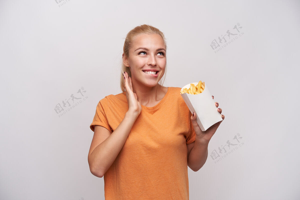 天然一个快乐的年轻可爱的金发女人的肖像 随意的发型咬着她的下颚 预感她正在吃薯条 在白色的背景下愉快地微笑勃艮第摆姿势饮食