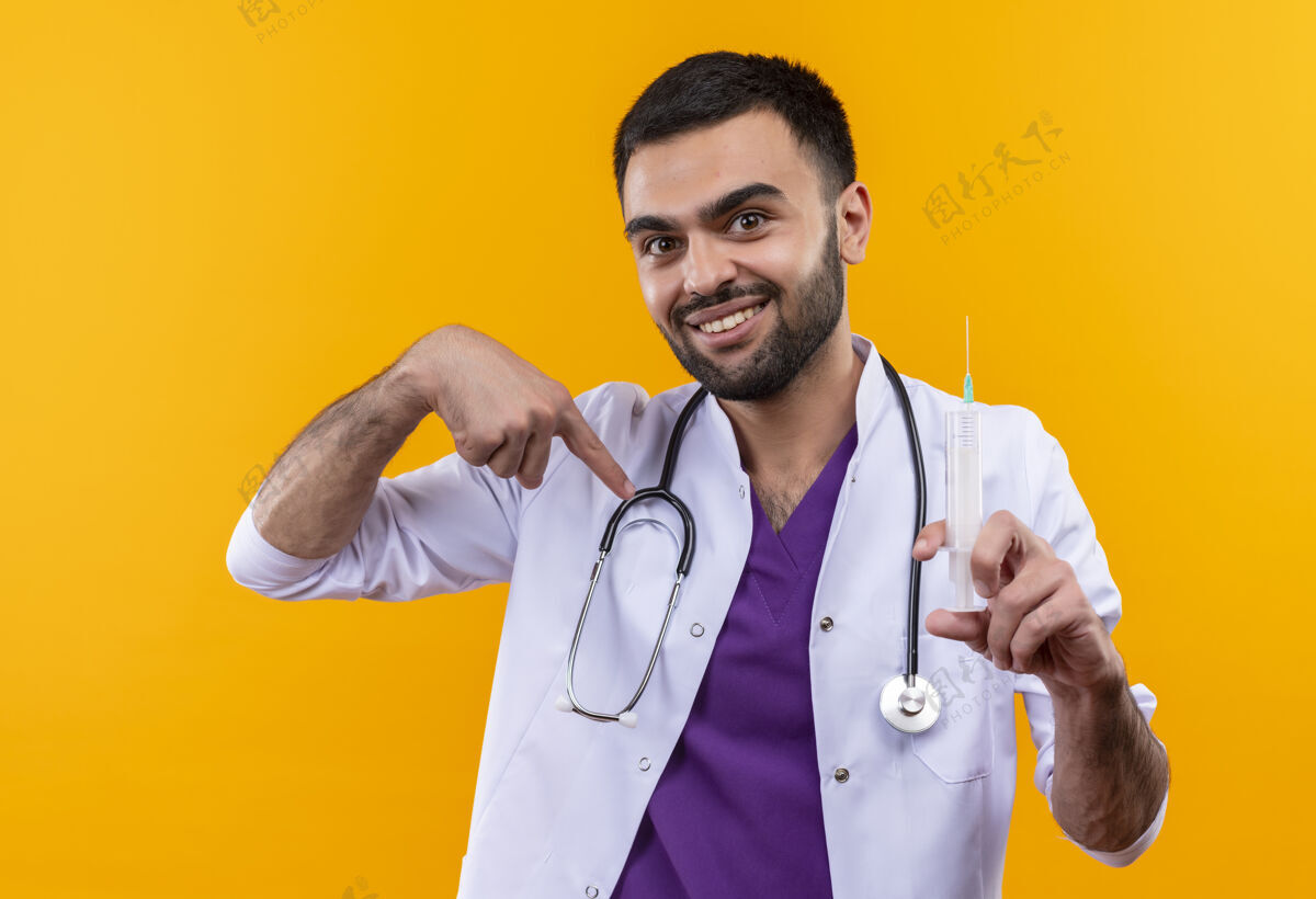 自己微笑的年轻男医生穿着听诊器医用长袍拿着注射器指着孤立的黄色背景上的自己听诊器长袍黄色