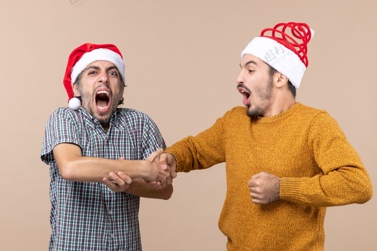 视图正面图：两个戴着圣诞帽的家伙在米色孤立的背景下痛苦地握手和喊叫肖像男性男人