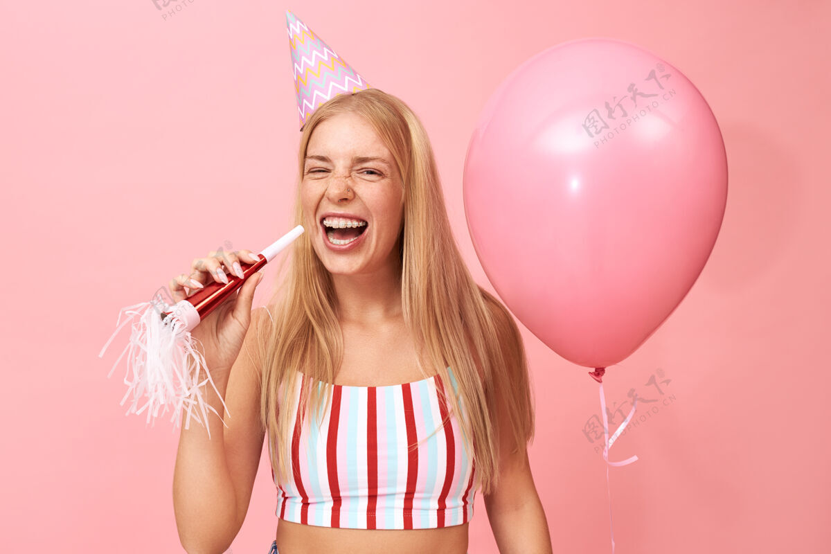 人美丽可爱的少女与松散的金发和背带的照片摆在粉红色与党吹风机和氦气球 笑着张大嘴巴女士庆祝手持