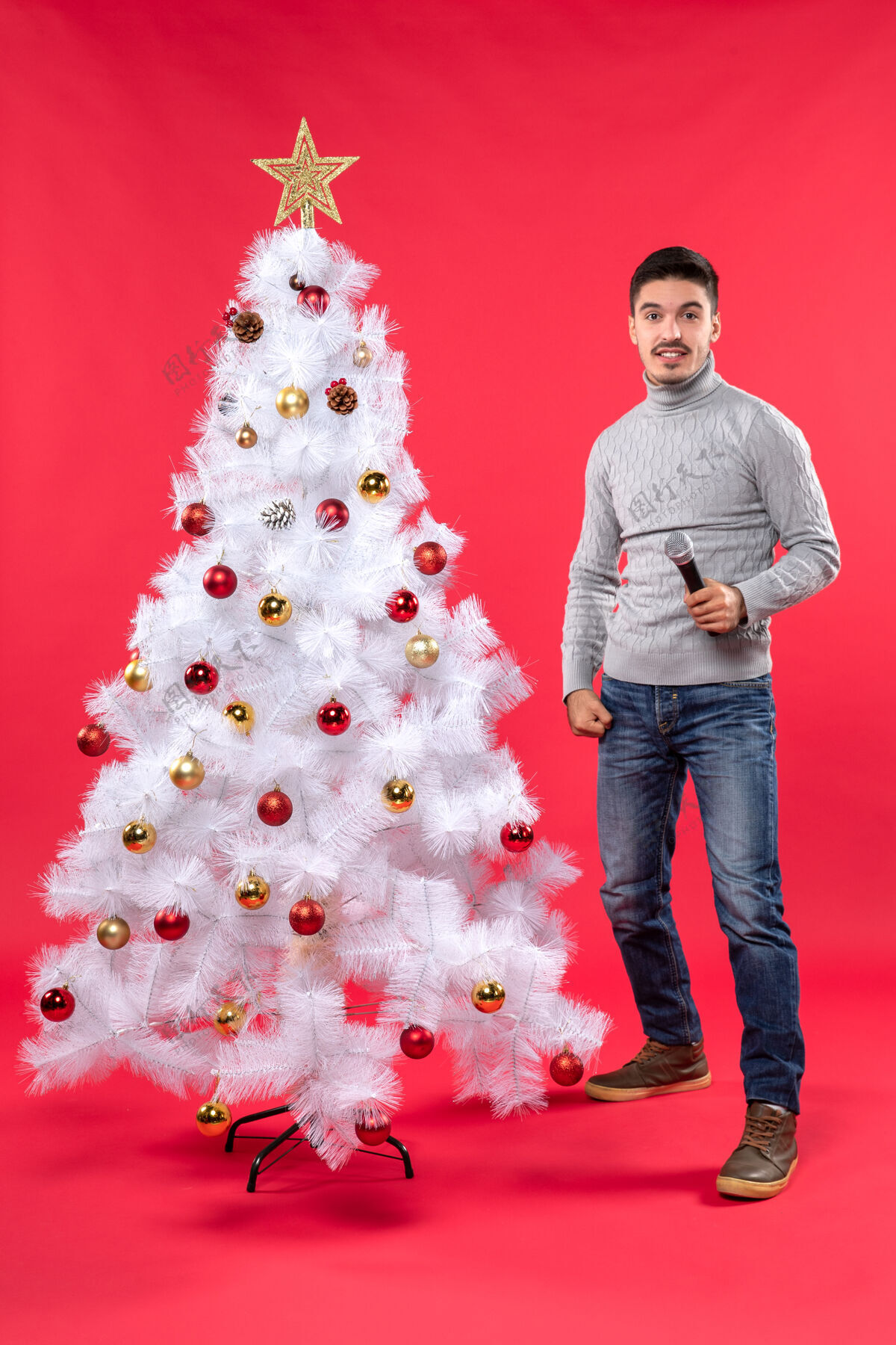 红色圣诞气氛 自信的家伙穿着牛仔裤站在装饰好的圣诞树旁 拿着麦克风 表情惊讶心情圣诞自信的男人