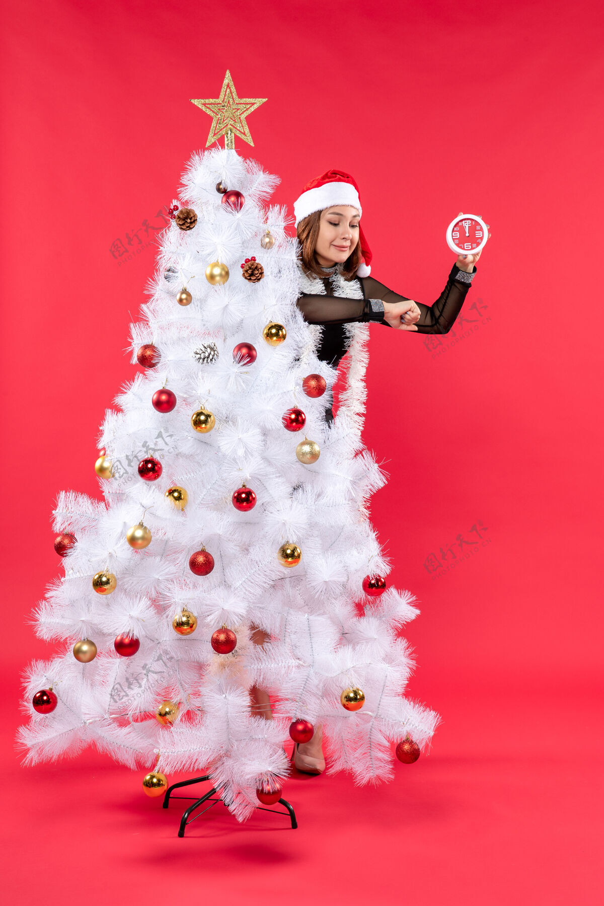 时间一个年轻漂亮的女孩 穿着黑色的裙子 戴着圣诞老人的帽子 躲在圣诞树后面 手里拿着时钟计时庆祝侧装饰