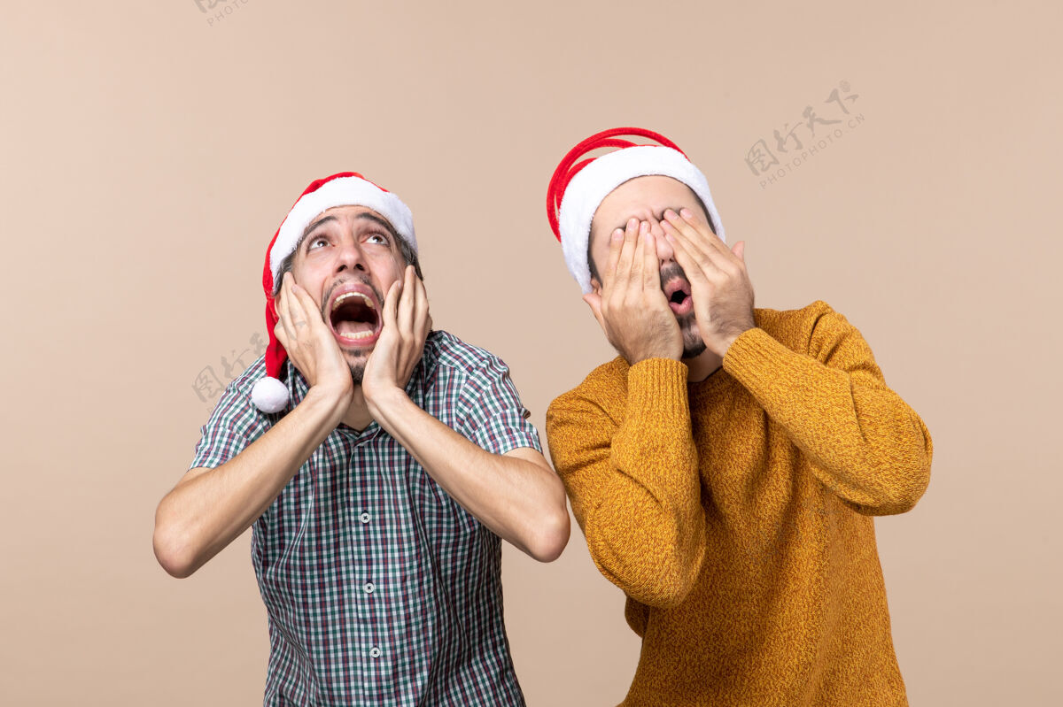 圣诞老人前视图两个戴着圣诞帽的吓坏了的家伙一个捂着耳朵 另一个在米色隔离背景上把脸埋起来帽子视图伙计们