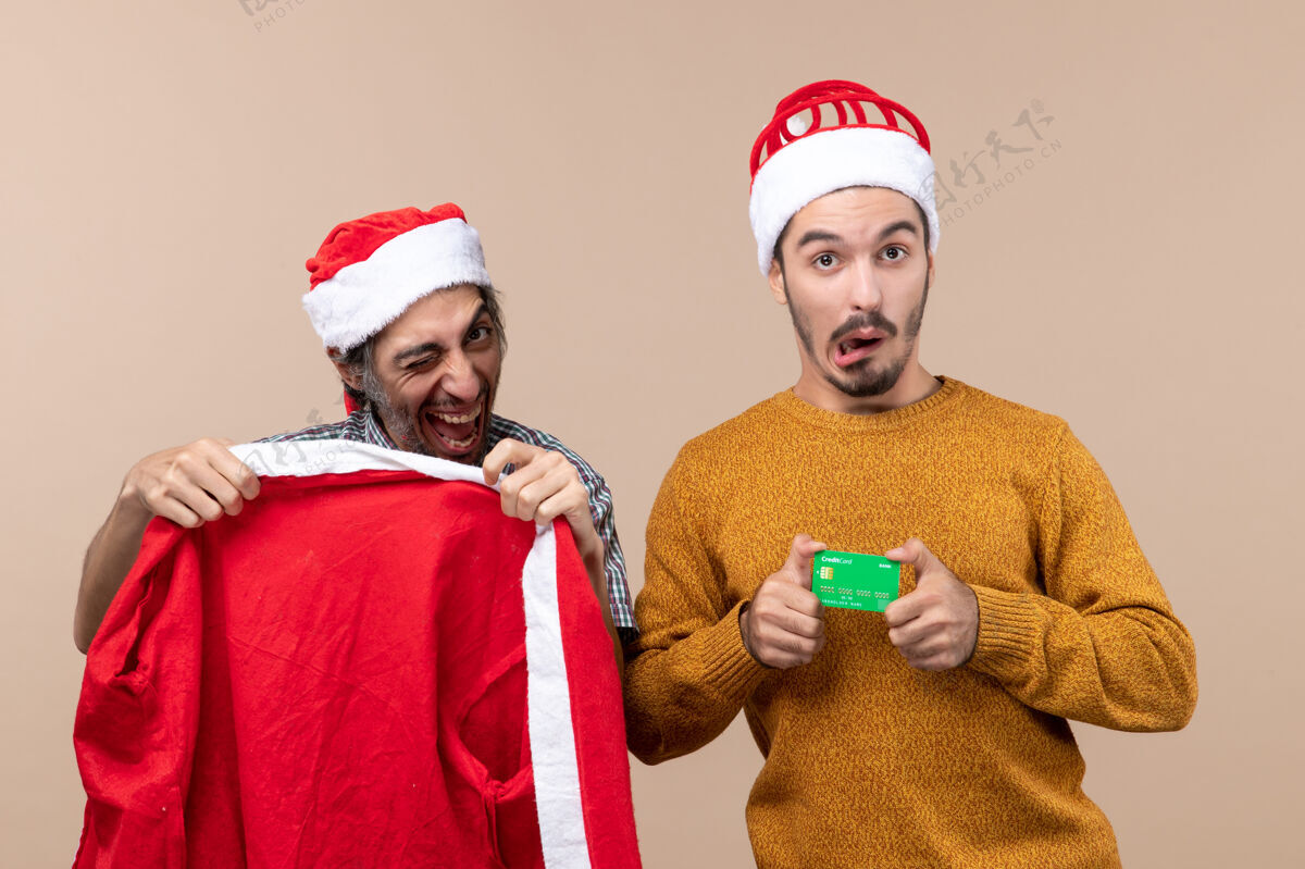 外套前视图两个家伙一个拿着圣诞老人的外套 另一个拿着信用卡 在米色孤立的背景下看着摄像机信用卡帽子男性