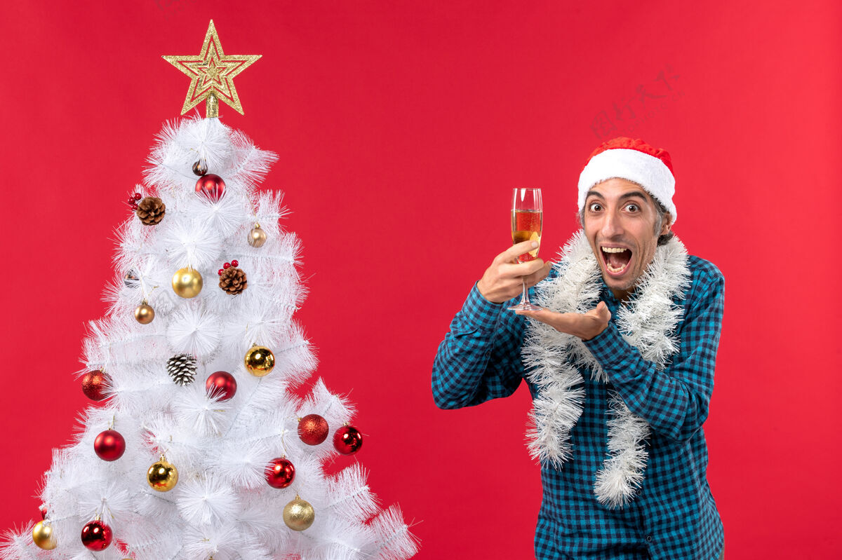 圣诞老人圣诞节心情与情绪愉快有趣的年轻人与圣诞老人帽子在一个蓝色条纹衬衫拿着一杯葡萄酒靠近圣诞树圣诞老人男人新年前夜