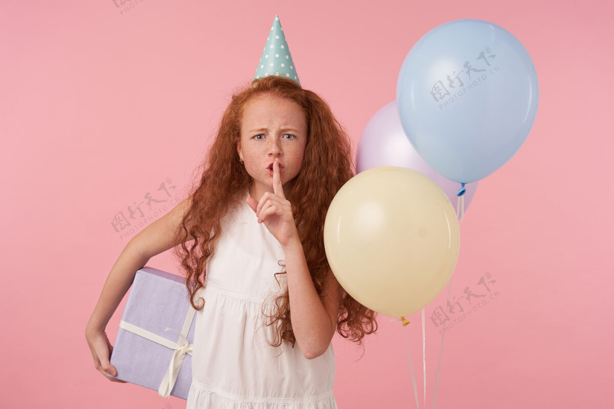 女孩严肃的小女孩 一头浓密的卷发 穿着节日的衣服 站在粉色的背景和彩色的气球上 手里拿着礼品盒 举起食指 要求保持沉默食指节日欧洲