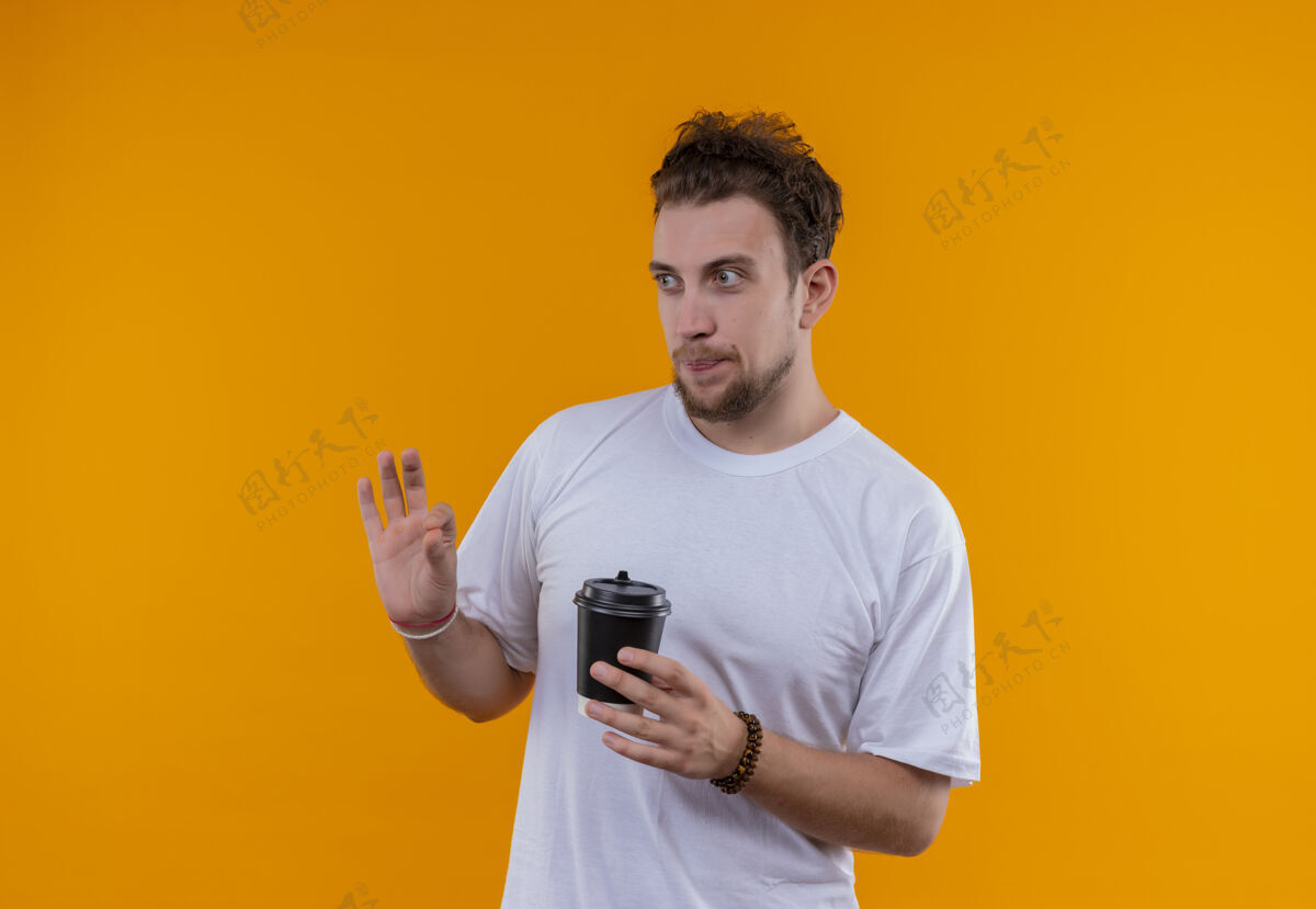 白色看着身边的年轻人穿着白色t恤 手里拿着一杯咖啡 在孤立的橙色背景上显示出良好的姿态好的年轻T恤