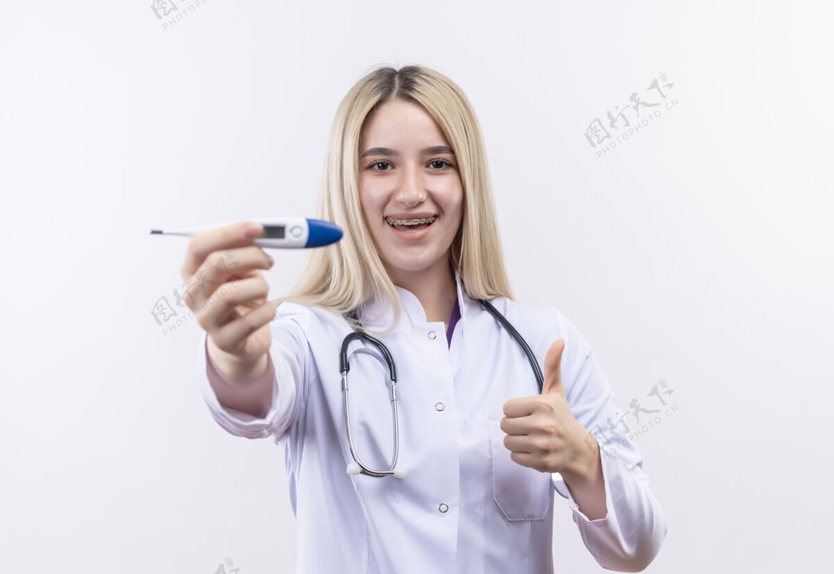抱着微笑的医生年轻的金发女孩 戴着听诊器和医用长袍 戴着牙套 拿着温度计对着摄像机 在孤立的白色背景上竖起大拇指金发医疗听诊器