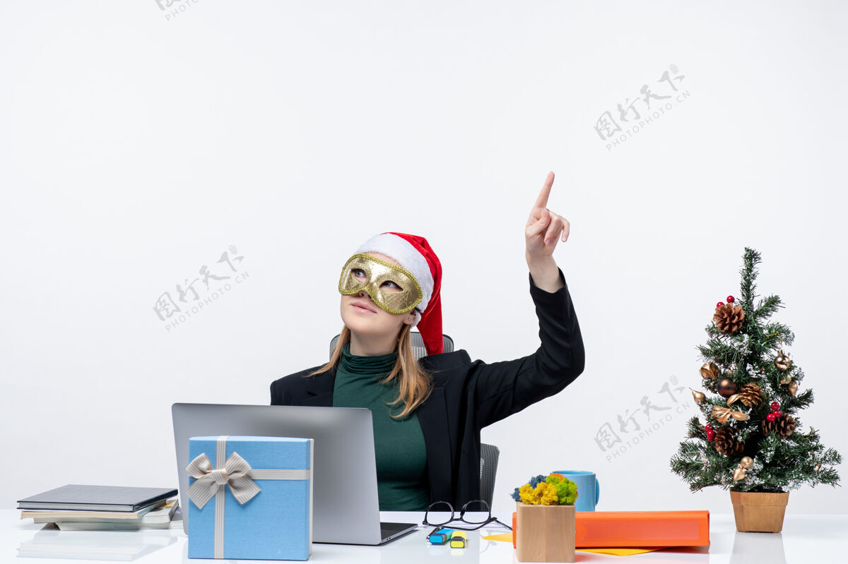 圣诞老人圣诞节心情与兴奋积极的年轻女子戴着圣诞老人的帽子 戴着面具坐在一张桌子上指着上面的白色背景坐着笔记本兴奋
