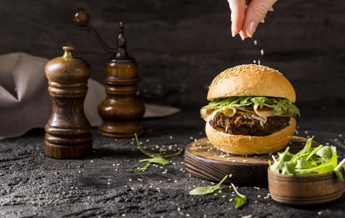 水平正面图：用手在牛肉汉堡上浇芝麻 配沙拉和培根汉堡包配料膳食