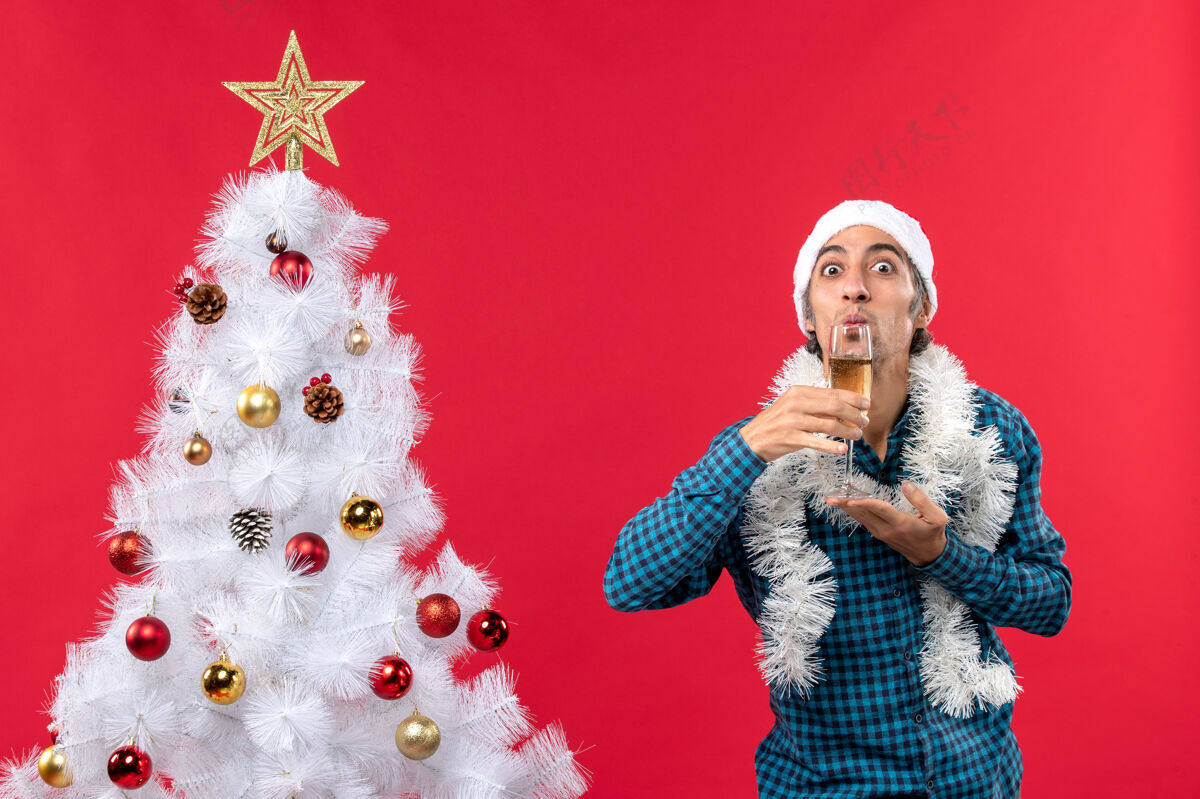 红酒圣诞节心情与情绪激动的年轻人圣诞老人帽子在一个蓝色的衬衫剥离举行和喝一杯葡萄酒附近圣诞树男人年轻新的