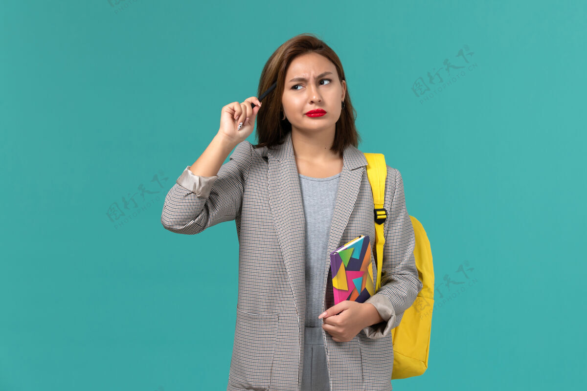 思考穿着灰色夹克 背着黄色背包 拿着文案 拿着钢笔思考的女学生在蓝色墙上的正视图保持正面字帖