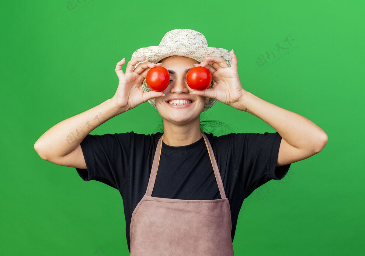 西红柿年轻漂亮的女园丁围着围裙 戴着帽子 手里拿着西红柿 眼睛蒙着 脸上带着微笑站着帽子绿色