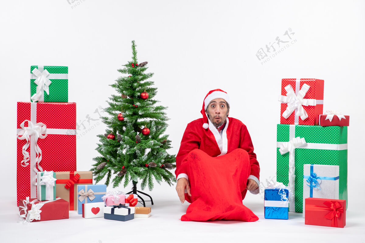 圣诞震惊的年轻人打扮成圣诞老人与礼物和装饰圣诞树坐在地上的白色背景礼物圣诞树坐