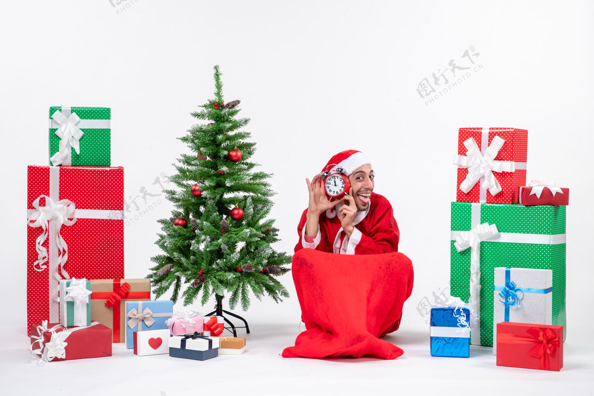 礼物情绪激动的快乐圣诞老人伸出舌头坐在地上 在礼物和白色背景上装饰圣诞树的旁边展示时钟礼物地盒子