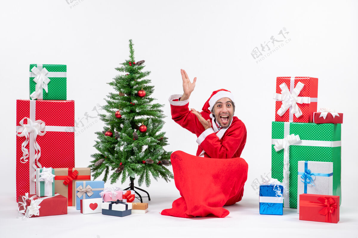 坐着有趣的年轻人打扮成圣诞老人与礼物和装饰圣诞树坐在地上指着上面的白色背景指向礼物礼物