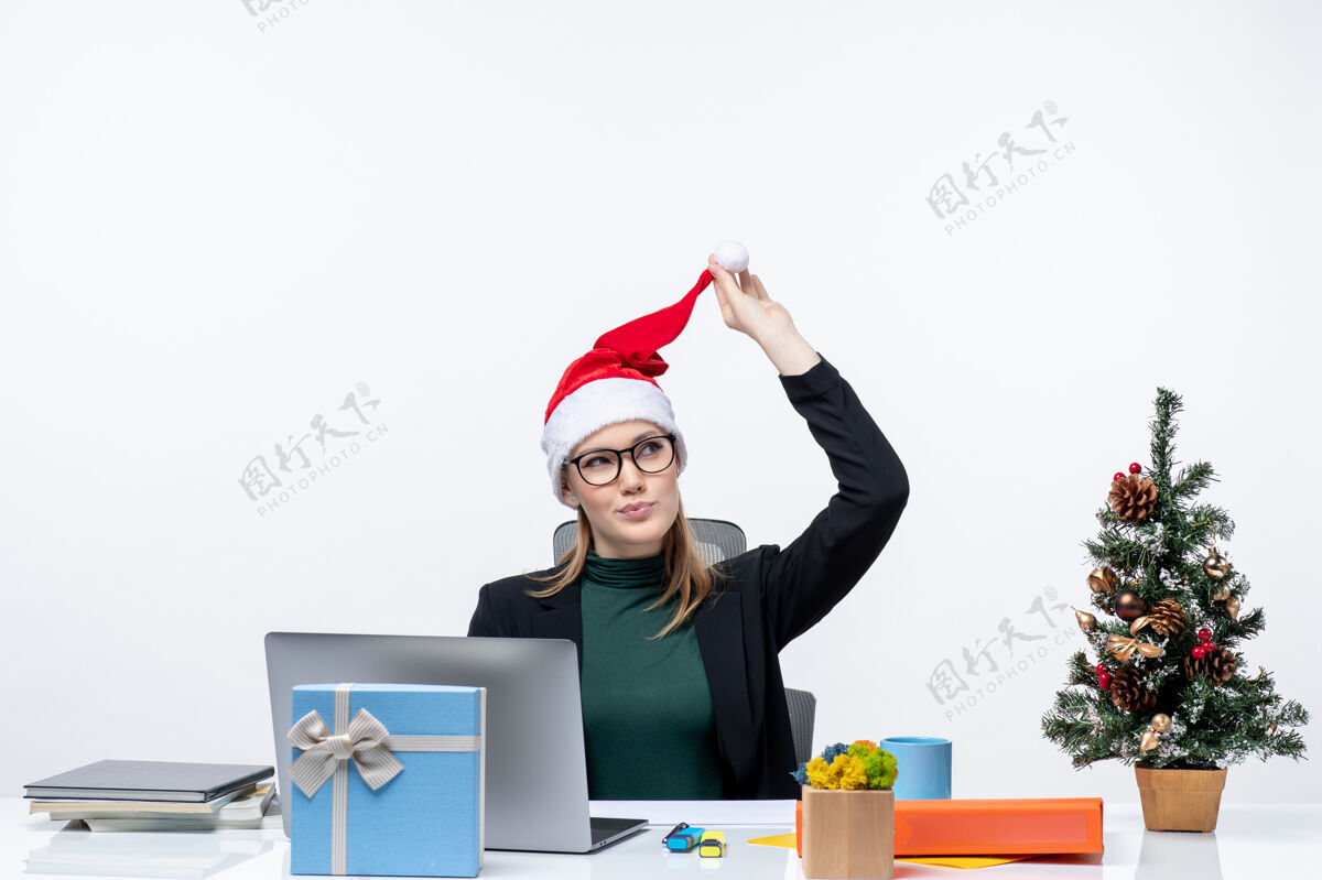 积极的一个积极的女商人正拿着圣诞老人的帽子坐在一张桌子旁 桌子上放着圣诞树和礼物 专注于白色背景的东西坐着帽子专注
