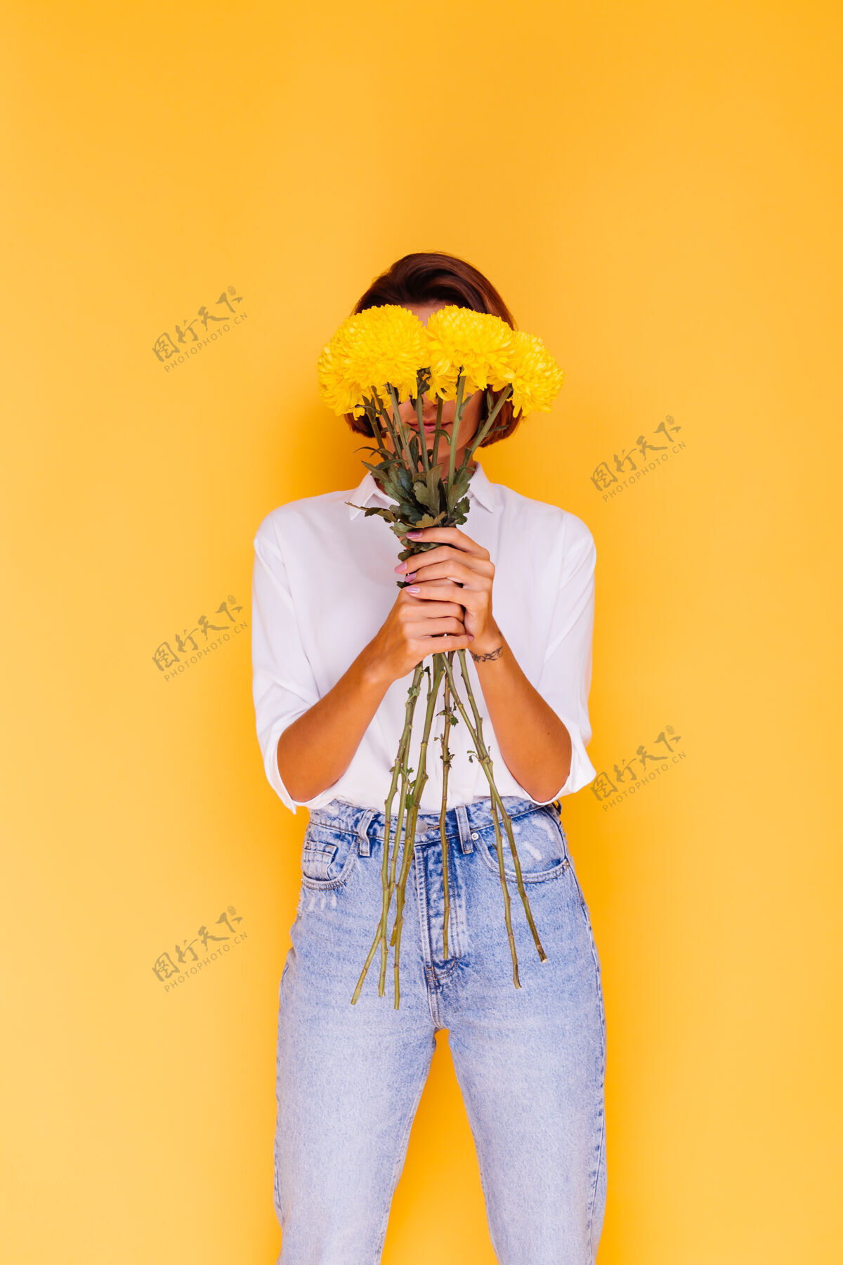 年轻摄影棚拍摄的黄色背景快乐的白人妇女短发穿着休闲服白衬衫和牛仔裤手持一束黄色紫苑气味积极可爱