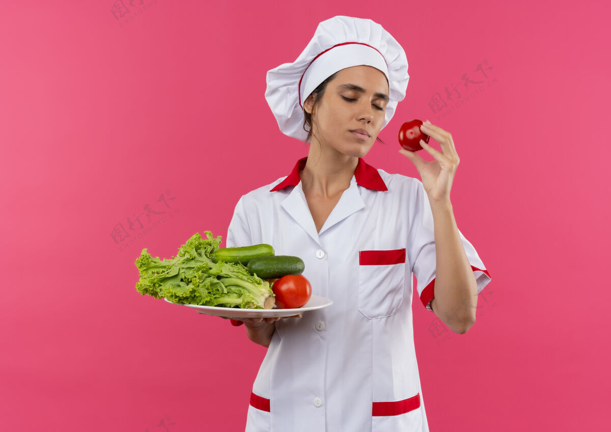 戴着年轻的女厨师闭着眼睛 穿着厨师制服 拿着蔬菜放在盘子里 留着复印空间厨师盘子蔬菜
