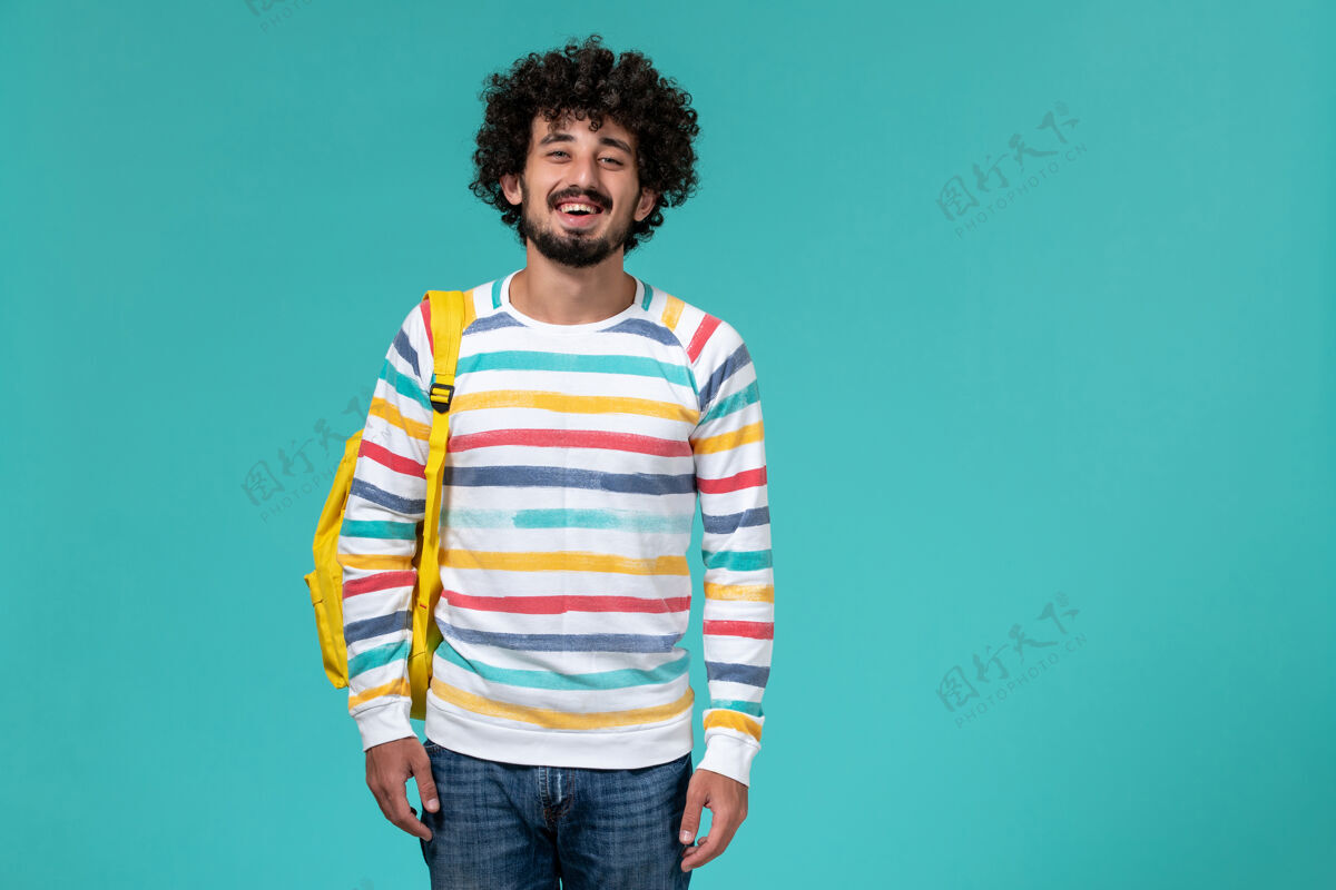 大学身着彩色条纹衬衫 背着黄色背包的男学生在蓝色墙上的正视图大学学生学校