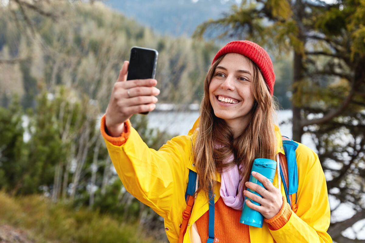 湖泊迷人的年轻女性游客在智能手机上自拍肖像 喝热咖啡或热水瓶茶采取风景自拍