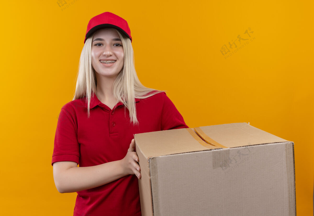 支撑微笑的小女孩穿着红色t恤 戴着帽子 戴着牙套 在孤立的橙色背景下拿着盒子帽子盒子橙色