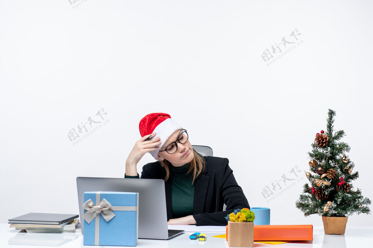 圣诞树一个疲惫的金发女人 戴着圣诞老人的帽子 坐在一张桌子旁 桌子上放着一棵圣诞树和一份白色背景的礼物礼物圣诞老人桌子