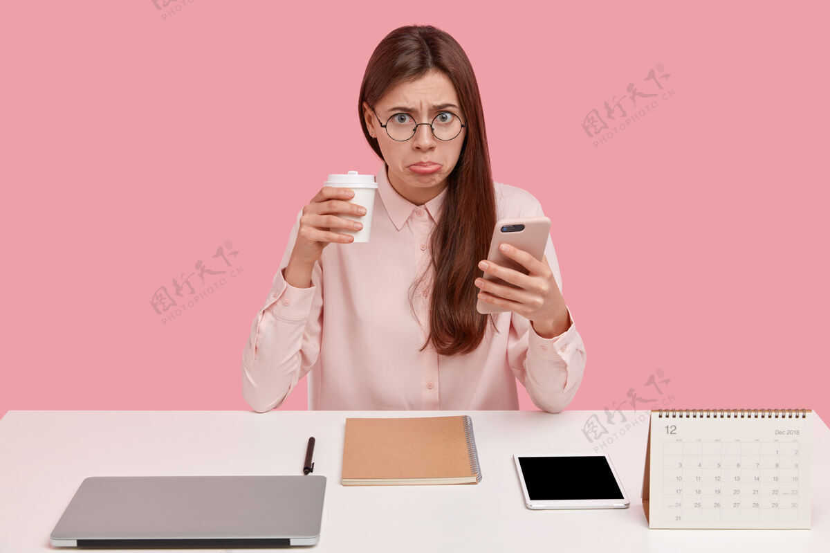 衬衫心烦的黑发女办公室完美主义者有不开心的表情 喝外卖咖啡 拿手机 戴圆眼镜自由职业者笔记本电脑