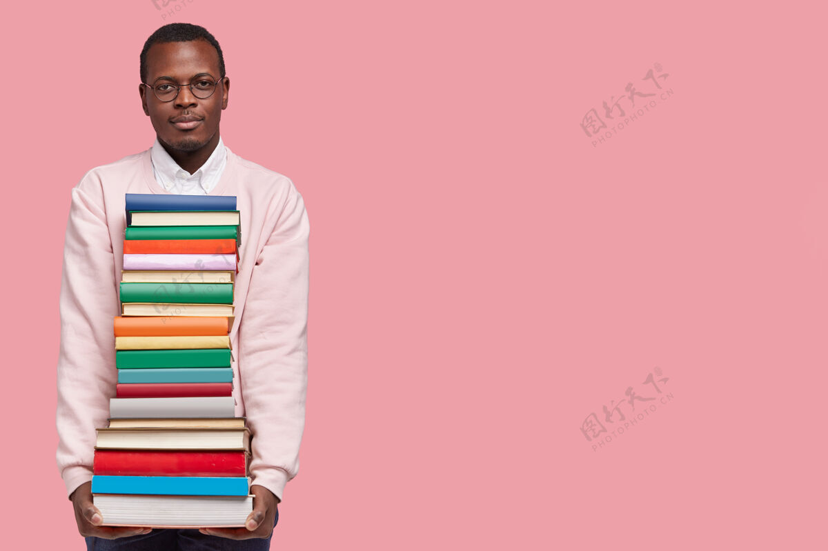 教育严肃的黑皮肤男学生的摄影棚镜头带着一大堆书 阅读和补习入学考试大学眼镜眼镜