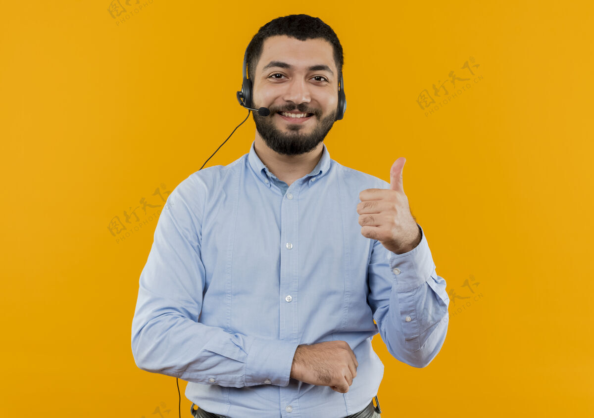 向上一个留着胡子的年轻人 穿着蓝色衬衫 戴着耳机 拿着麦克风 微笑着竖起大拇指看年轻耳机
