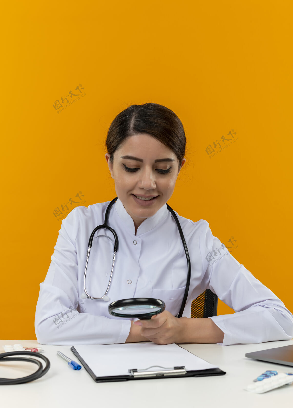 医疗微笑的年轻女医生穿着医用长袍 手持听诊器 坐在办公桌旁 拿着医疗工具 用放大镜在黄色背景上看剪贴板女工具听诊器