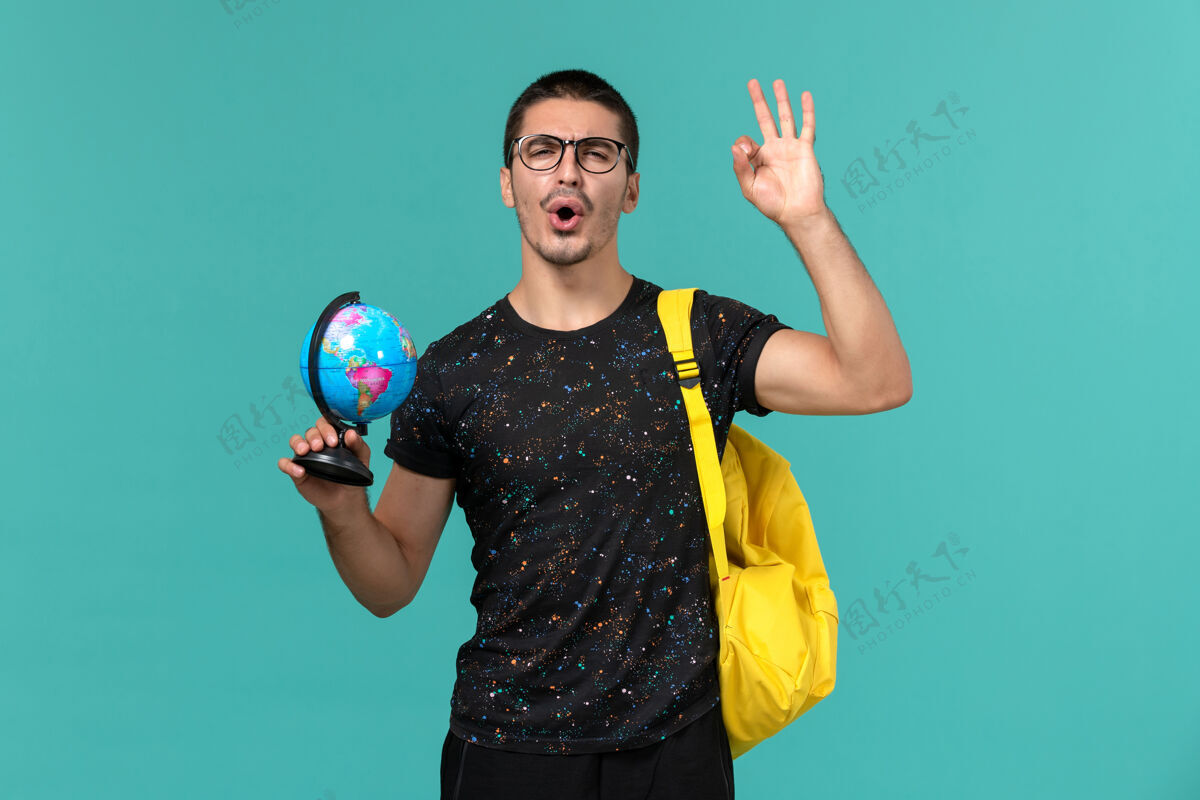 马拉卡身穿深色t恤黄色背包的男学生正面图蓝色墙上拿着小地球仪男学生大学小