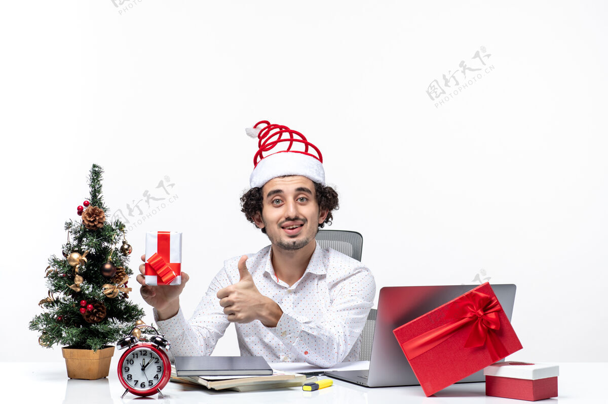 帽子圣诞节心情与快乐情绪与年轻的商人与圣诞老人帽子和提高他的礼物 使白色背景的ok手势圣诞老人礼物年轻