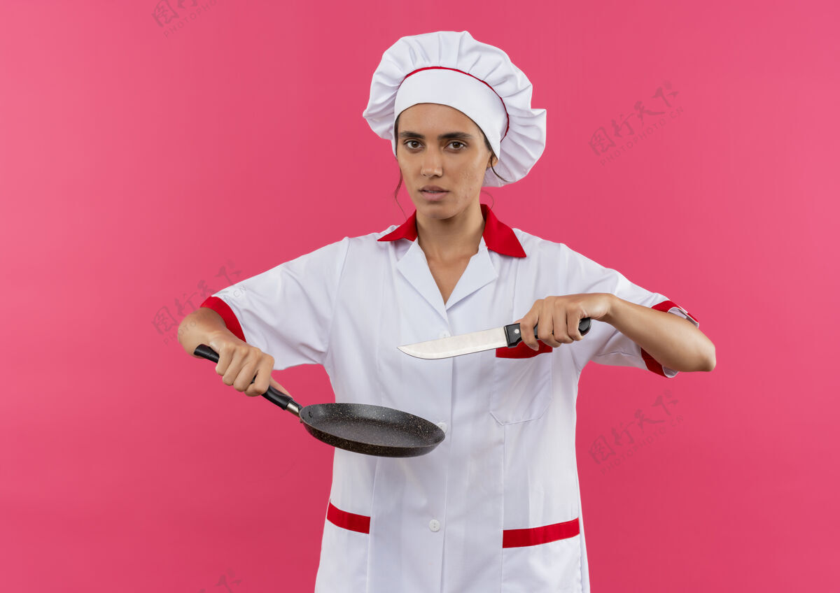 平底锅年轻的女厨师穿着厨师制服 拿着煎锅和刀站在隔离的粉色墙壁上 还有复印空间厨师拿着看