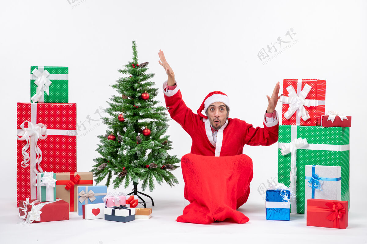 礼物困惑兴奋的年轻人打扮成圣诞老人 拿着礼物和装饰好的圣诞树坐在白色背景的地上礼物圣诞老人困惑