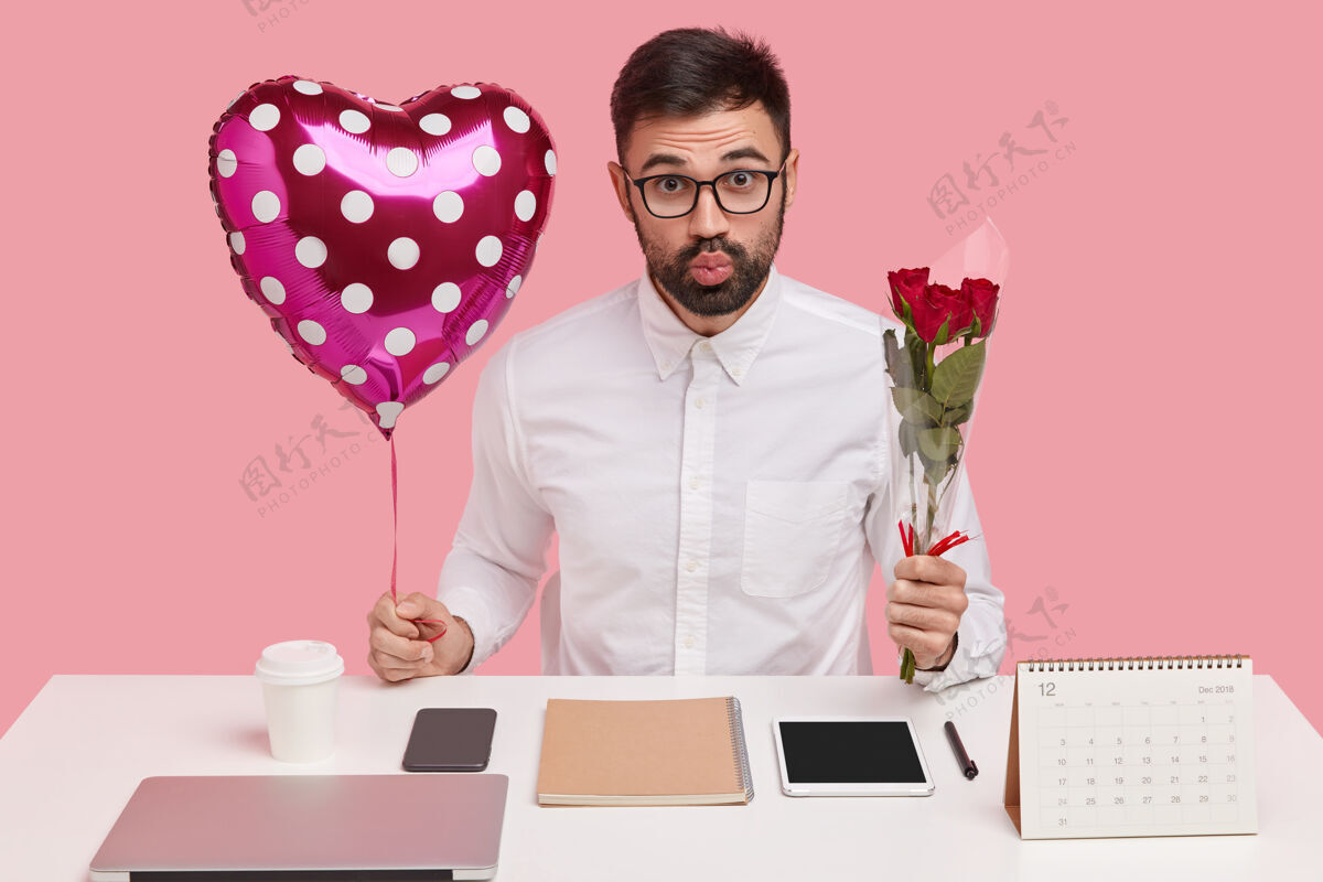 衬衫室内拍摄的帅哥没刮胡子的男人撅着嘴唇 带着情人节和花束 在办公室里有浪漫的关系 在粉色的工作室墙上摆姿势男性饮料饮料