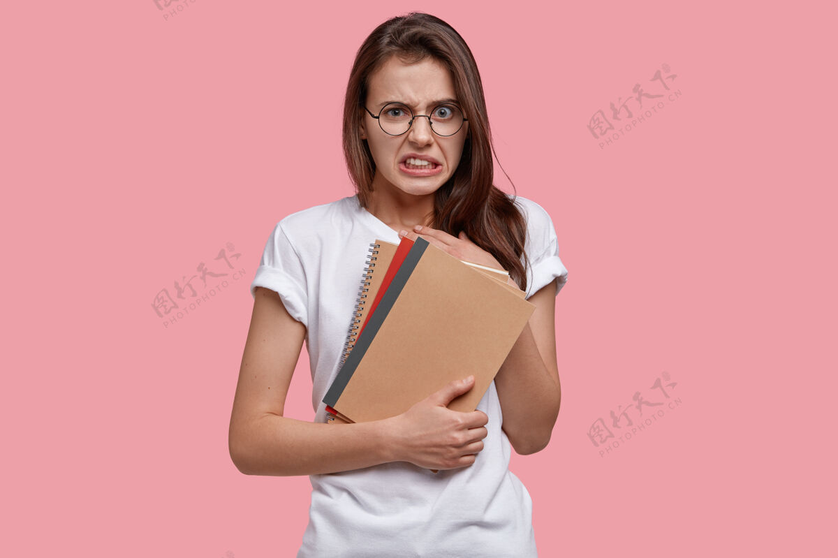 学习不高兴的大学生女士有不高兴的表情 咬紧牙关 拿着螺旋笔记本消极眼镜笔记本