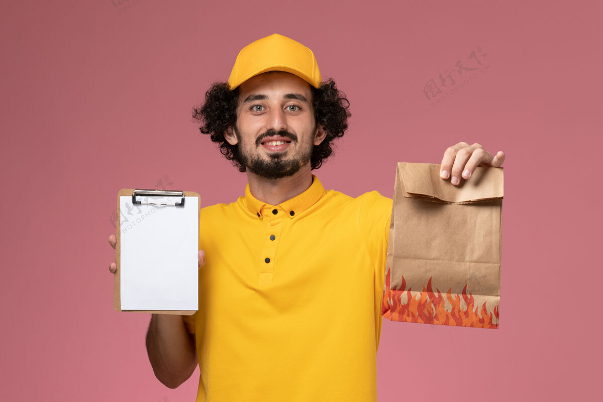 黄色正面图身穿黄色制服的男快递员拿着食品包和笔记本 放在浅粉色的墙上正面持有男快递