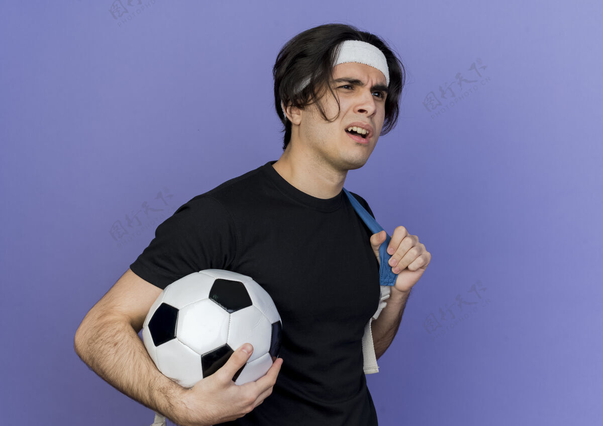 一边年轻的运动型男子穿着运动服 戴着头巾 背着背包 手里拿着足球 一边看一边又困惑又不高兴年轻运动足球