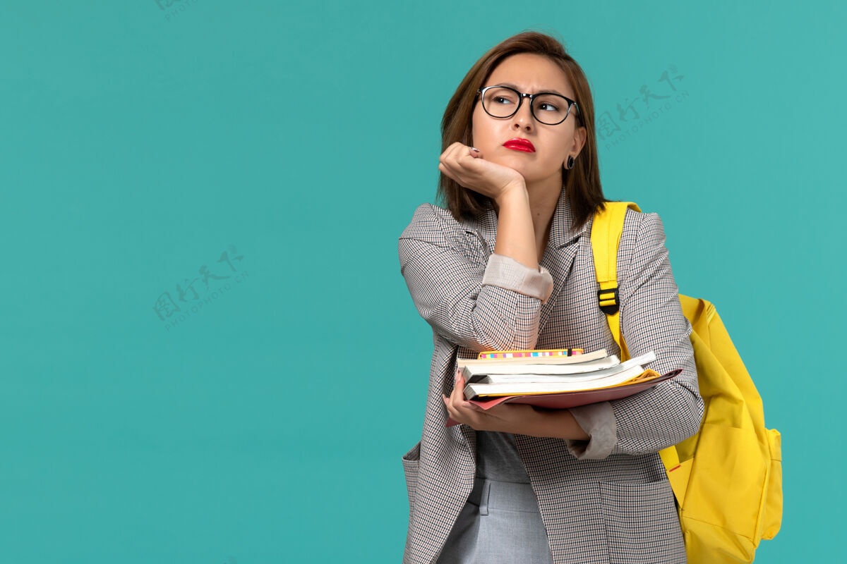黑发穿着灰色夹克的女学生的正面图黄色背包拿着书在蓝色的墙上思考模型大学成人